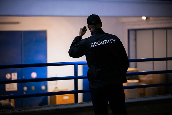 A security guard in uniform, holding a flashlight, standing in front of a building at night.