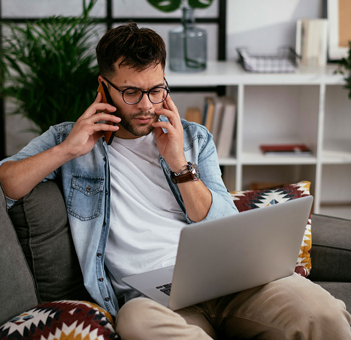 Man in denim casually multitasking on phone and laptop, pondering over Canadian questions, seated on a colorful couch.