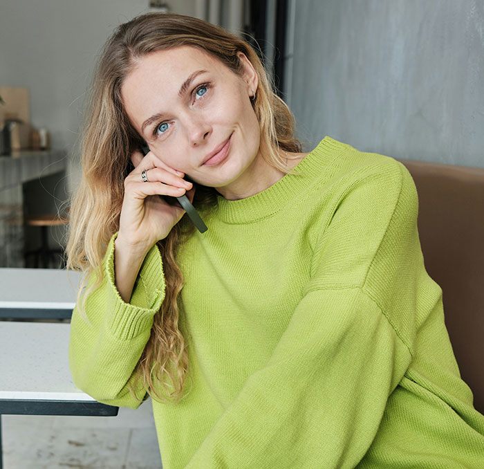 Woman in a green sweater sits at a cafe table, smiling and holding a pen, contemplating Canadian language questions.