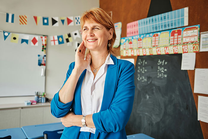 A smiling woman in a classroom, wearing a blue blazer, with educational decor and a chalkboard in the background.