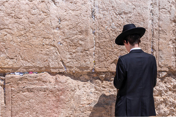 A person in traditional attire facing a historic stone wall, partially shadowed, suggesting reflection or prayer.