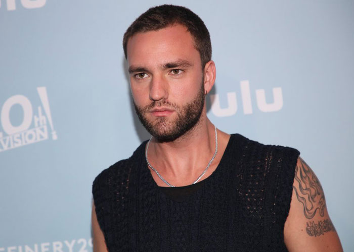 Man from Tell Me Lies cast in a black sleeveless shirt at a Hulu event, with a tattoo on his left arm.