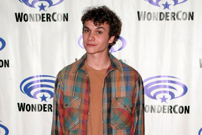 Cast member of "Tell Me Lies" at WonderCon, wearing a plaid shirt, posing in front of event backdrop.