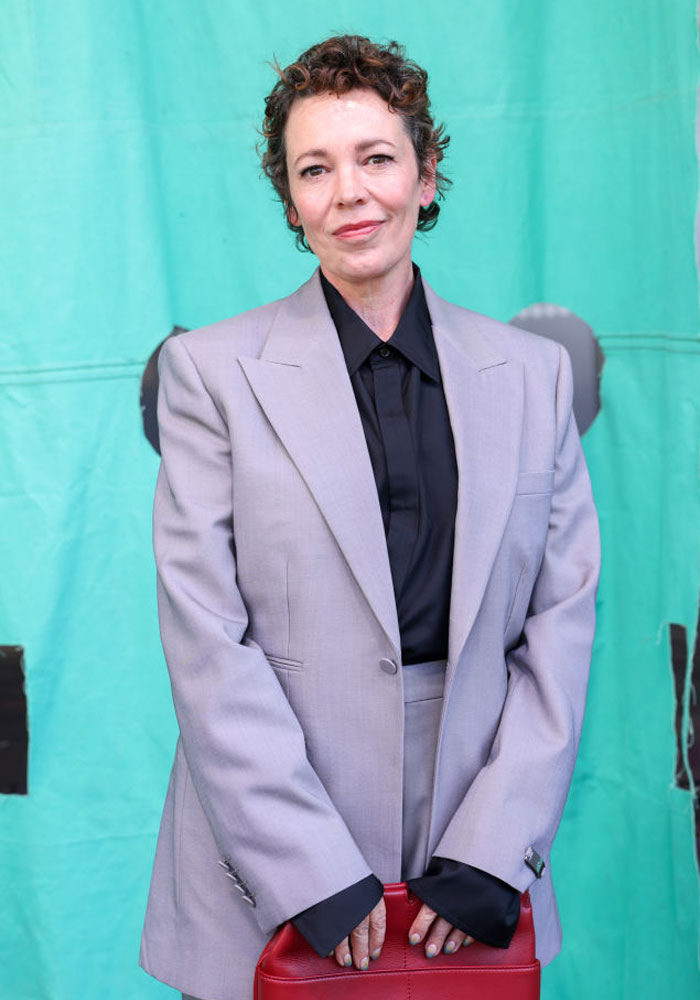 Olivia Colman in a gray suit, part of "The Bear" cast, poses against a teal backdrop, holding a red bag.