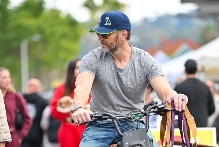 Joel McHale in gray shirt and cap riding a bike, representing stars from The Bear cast in a park setting.