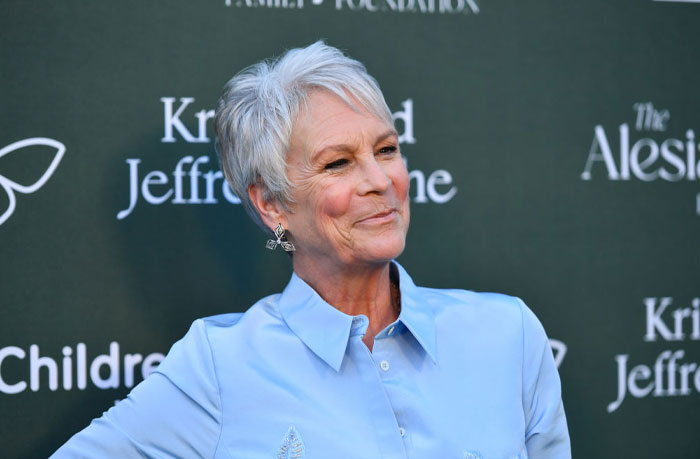 Jamie Lee Curtis smiling at an event, promoting The Bear cast, wearing a light blue shirt against a green backdrop.