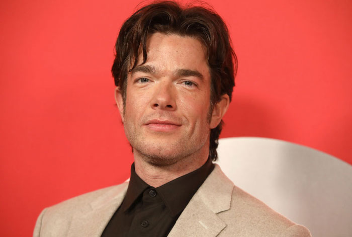 John Mulaney from The Bear Cast season 3 in a beige suit against a red backdrop.