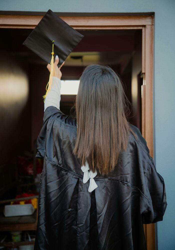 Graduate in black robe holding a cap, standing in a doorway.