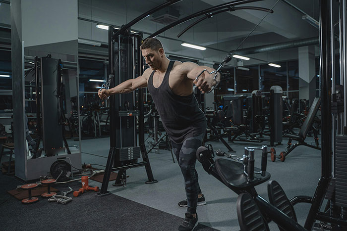 Man exercising in gym, wearing a black tank top, surrounded by fitness machines and weights, showcasing unique fitness routine.