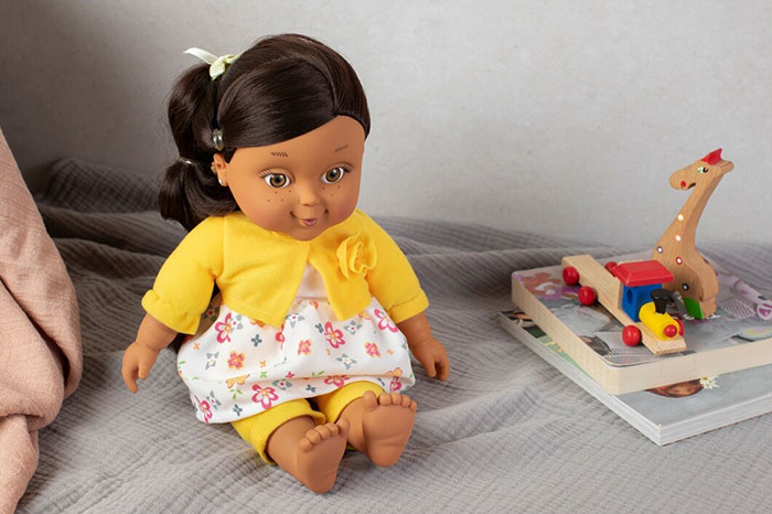 A doll in a floral dress and yellow cardigan, representing things people bought as adults denied in childhood, on a bed.