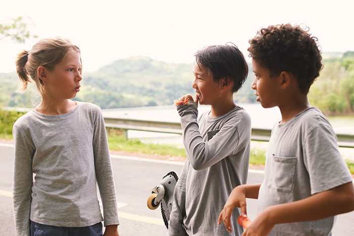 Three kids in casual clothes stand near a road, with one holding a skateboard, enjoying a conversation on a sunny day.