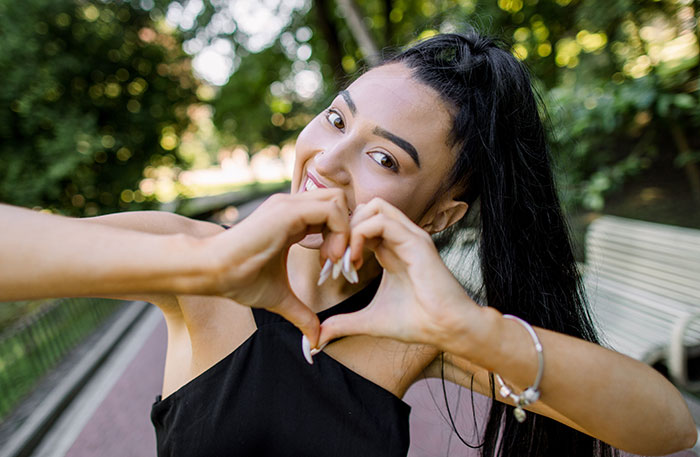 Person making a heart shape with hands, smiling in a park, highlighting romanticized moments.