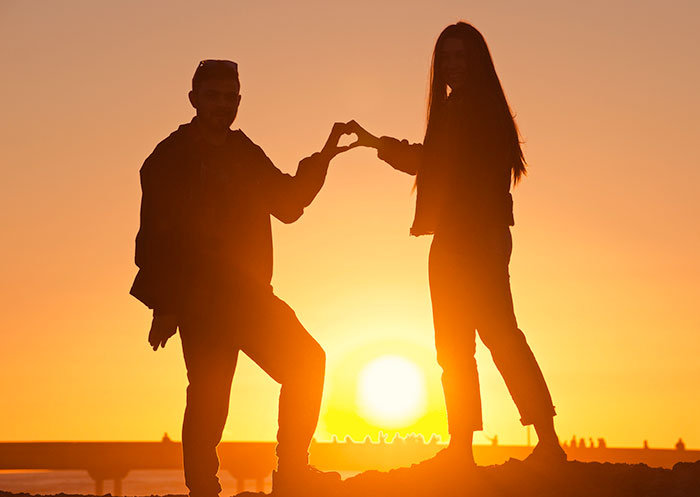 Silhouette of a couple forming a heart shape with hands against a sunset, highlighting romanticized moments.