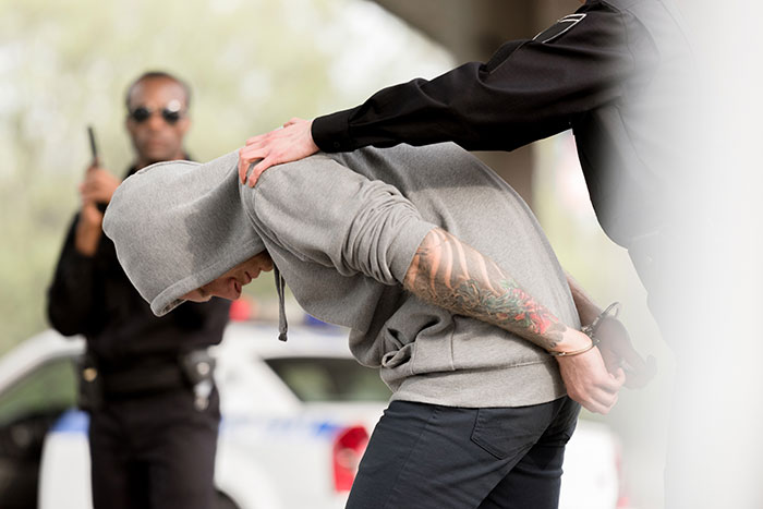 Person in hoodie being arrested by police, illustrating harsh reality versus romanticized perception.