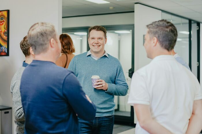 Group chatting in an office, one man holding a coffee cup, illustrating social interactions people secretly judge others for.