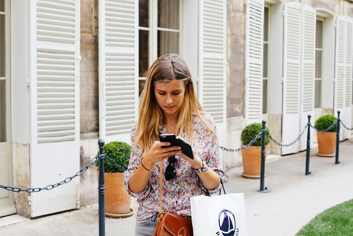 Woman walking outdoors, focused on her phone, holding a shopping bag; example of things people judge others for.