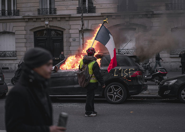 A burning car with a protester holding a French flag, illustrating terrible chaos in a city setting.