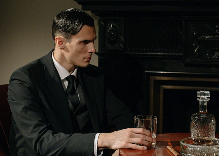 A man in a suit sitting by a table with a glass of whiskey, reflecting on terrible choices.