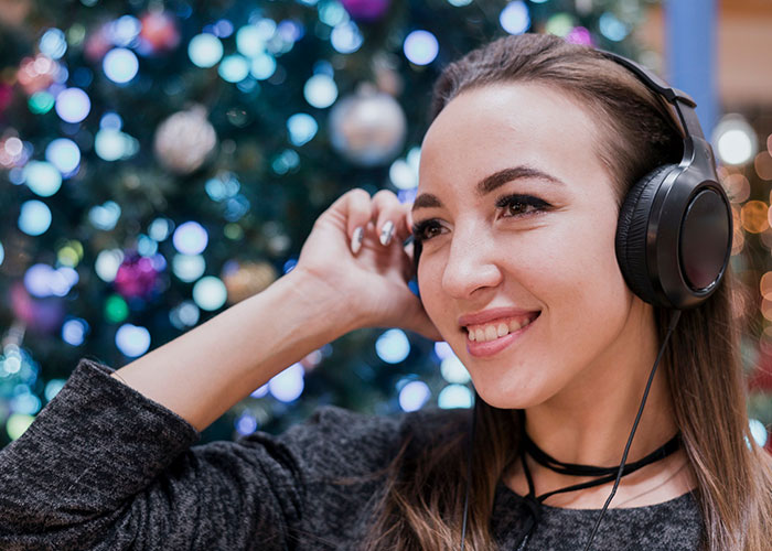 Woman enjoying music with headphones, smiling near decorated tree, capturing intriguing moment.