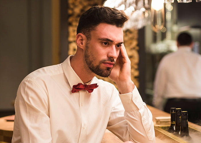 Man in a white shirt with a red bow tie sitting at a bar, appearing contemplative, representing intriguing facts discussed.