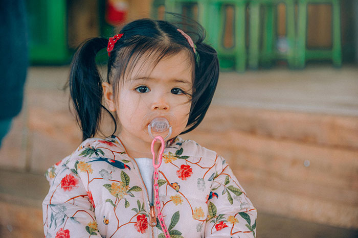 A young girl with pigtails and a pacifier, wearing a floral jacket, outdoors.