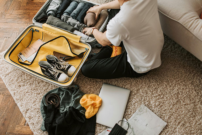 A person packing a suitcase with clothes and shoes on a carpeted floor.