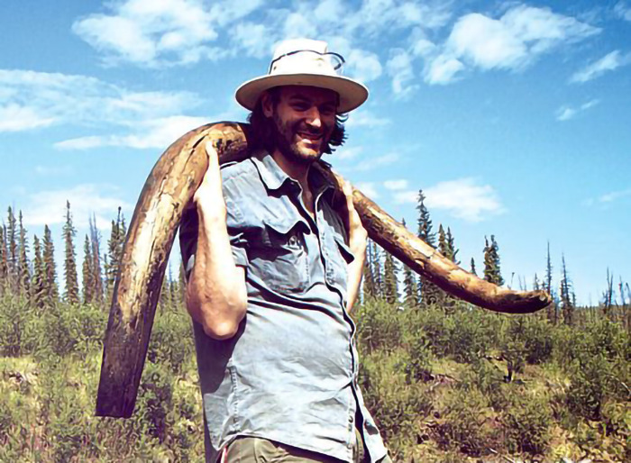 Man with woolly mammoth tusk in a forest setting.