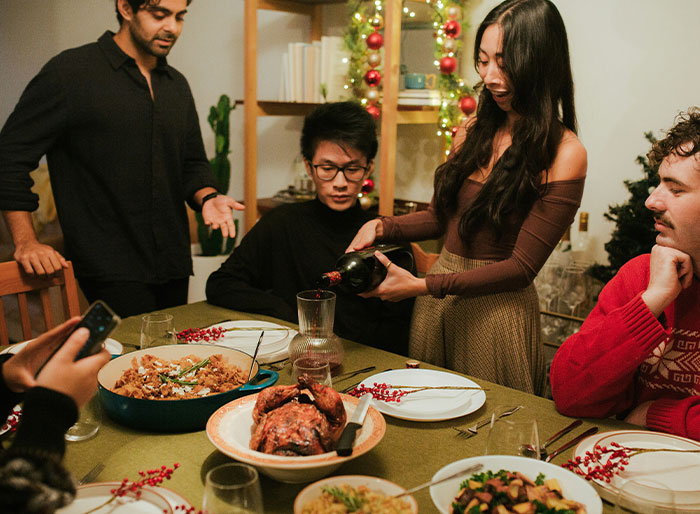 Family gathered around a festive dinner table with holiday decorations, a woman pouring wine.