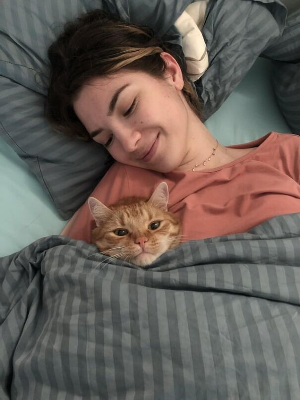 A person smiling with a ginger cat tucked in beside them under a grey-striped blanket.