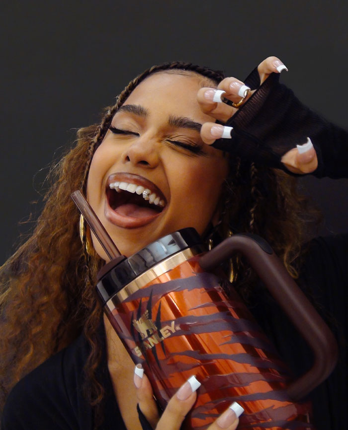 Tyla smiles brightly, holding a metallic tiger-striped tumbler, showing off her braces and white-tipped nails, radiating playful energy.