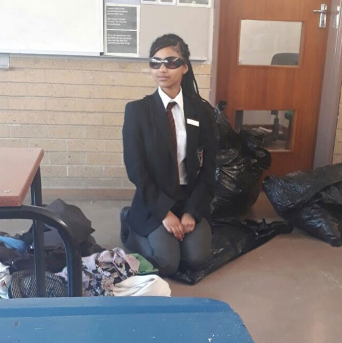  Tyla during her school years, wearing a formal uniform with a blazer, tie, and sunglasses, kneeling in a classroom near black garbage bags and folded clothes. The setting appears casual yet formal, with a classroom door and bulletin board in the background.