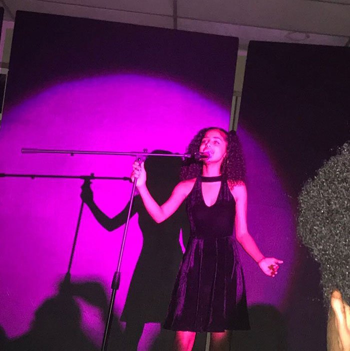 Tyla singing passionately on stage under a dramatic purple spotlight. She wears a black halter dress and holds a microphone, her shadow cast vividly behind her. The atmosphere suggests a live performance or talent showcase.