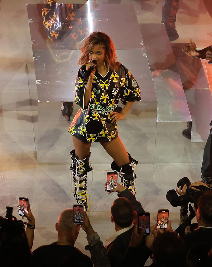 A dynamic performance shot of Tyla on stage, wearing a bold black and yellow patterned outfit paired with high lace-up boots. She sings confidently into a microphone, engaging with a captivated audience capturing the moment on their phones, while dramatic lighting highlights her presence on a reflective stage.