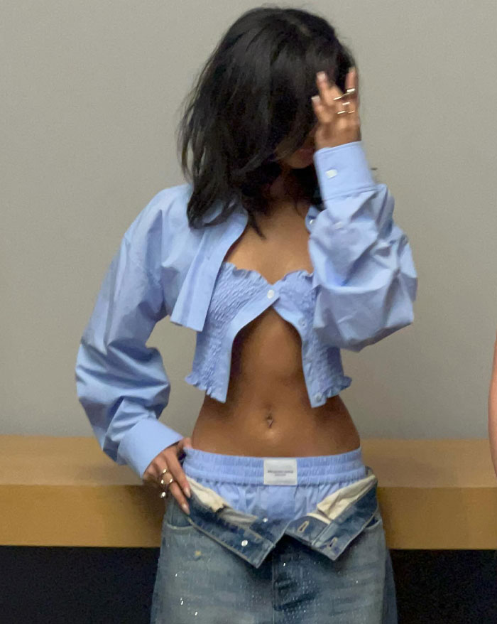 Tyla poses casually, wearing a cropped light blue shirt over a matching smocked bandeau and layered with oversized denim jeans styled low on the hips. She playfully obscures part of her face with her hand, accessorized with rings, creating a laid-back, edgy aesthetic.