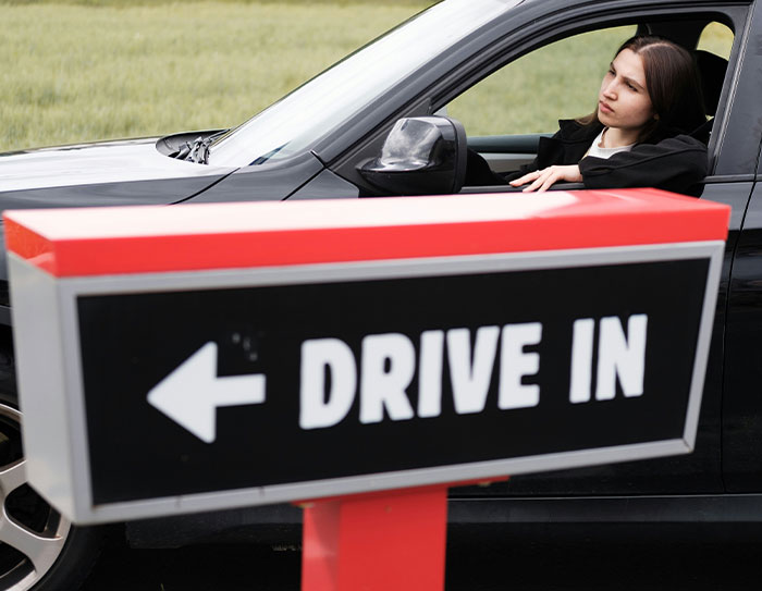 Person using a drive-in sign the wrong way, sitting in a black car and looking away.