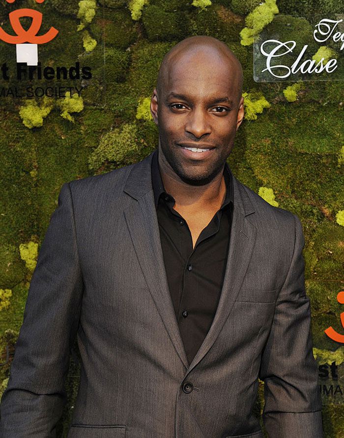 "Actor from Virgin River cast in a grey suit, smiling at an event with mossy green backdrop."