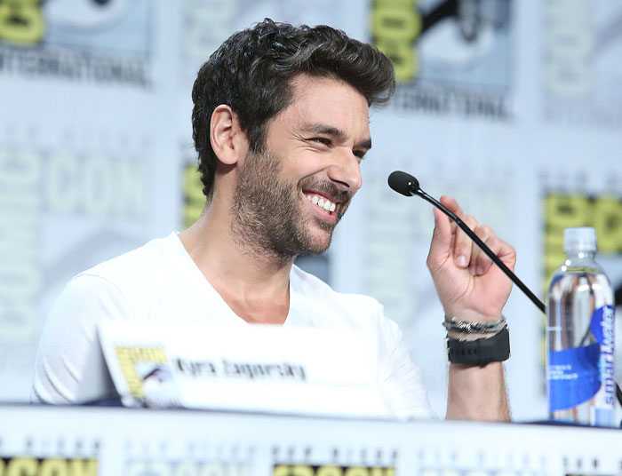 Smiling Virgin River cast member at a Comic-Con panel, wearing a white shirt, with a microphone and water bottle nearby.