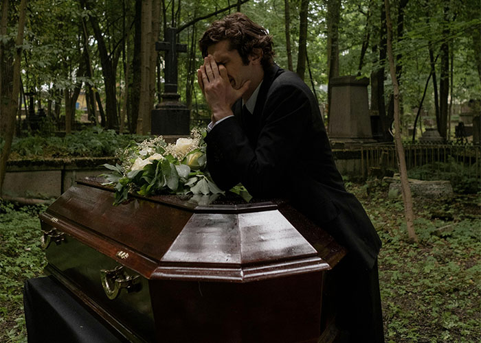 A man in a suit mourns at a coffin with flowers in a cemetery, evoking feelings of mystery and unanswered questions.