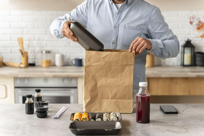 Man in a blue shirt unpacking sushi delivery on a kitchen counter, with soy sauce and juice.