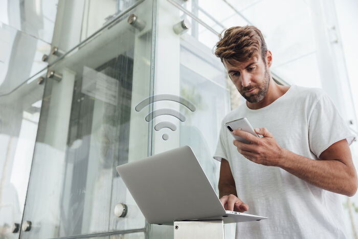 Man using laptop and phone in a glass building lobby, appearing focused and connected.