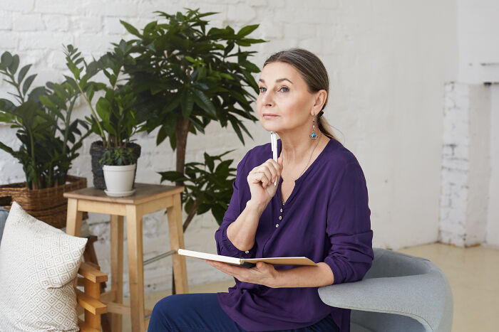 A woman in a purple shirt sitting in a chair, holding a pen and notebook, with houseplants in the background.