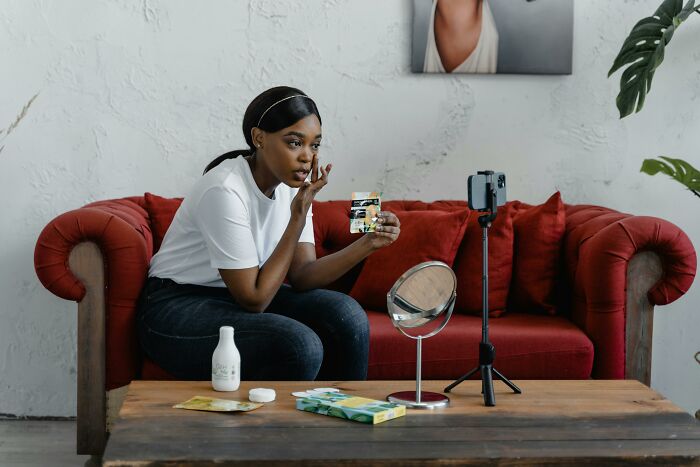 A woman sitting on a red sofa, applying makeup, recording herself with a phone.