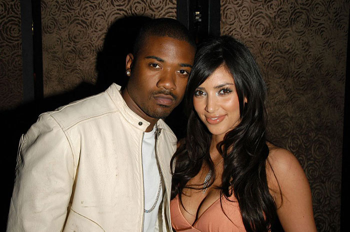 A photo of Kim Kardashian posing with a man in a light jacket at an event, both smiling slightly, with a patterned background.