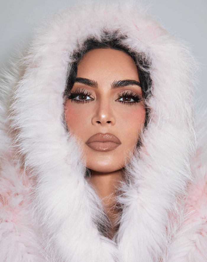 Kim Kardashian in a close-up portrait wearing a luxurious pale pink fur-lined hood, showcasing her glamorous makeup and glossy lips.