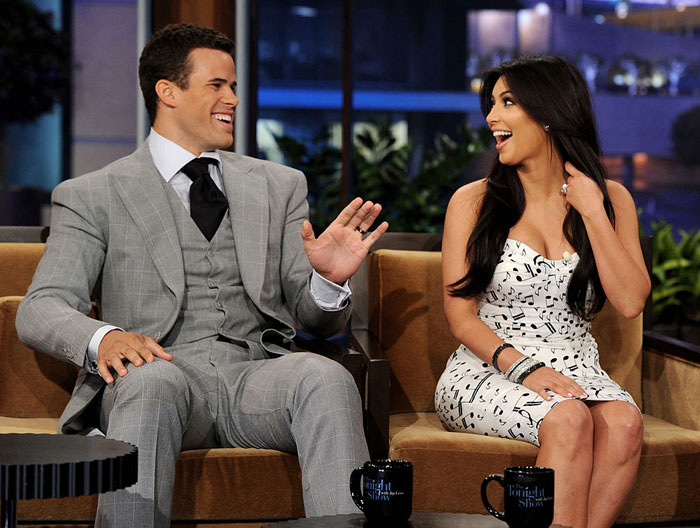 Kim Kardashian and a man in a gray suit sitting on a talk show couch, both laughing, with mugs on the table and a nighttime cityscape background.