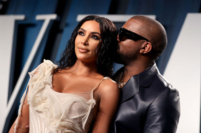 Kim Kardashian in a white dress standing beside a man in a leather jacket and sunglasses, with him leaning close to her, set against a blue backdrop.