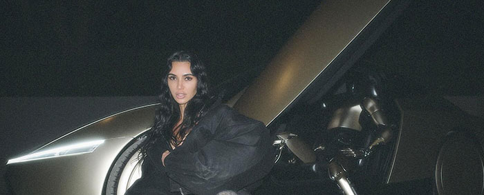 Kim Kardashian wearing a dark outfit, crouching in front of a futuristic gold car with its doors open at night, exuding a sleek and modern vibe.