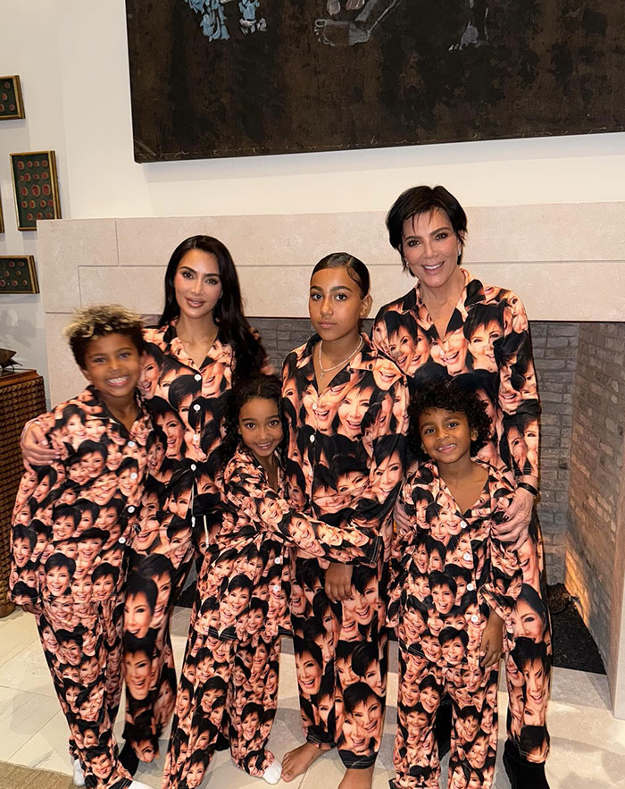 Kim Kardashian posing with family members, all wearing matching pajamas featuring a printed face design, standing in a cozy room with a fireplace.