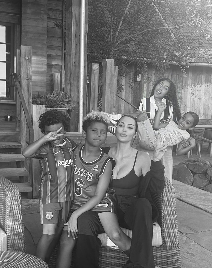 Kim Kardashian sitting outdoors with her children in a black-and-white photo, featuring a rustic wooden background and playful poses from the kids.