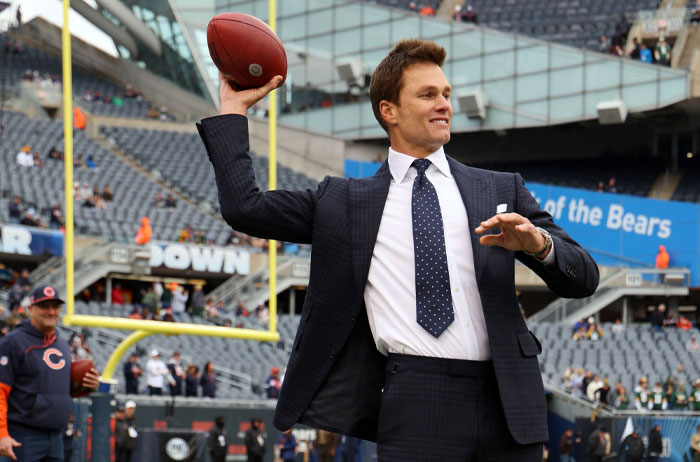 A man in a suit, identified as Tom Brady, smiles as he prepares to throw a football in a stadium, blending formality with sports.